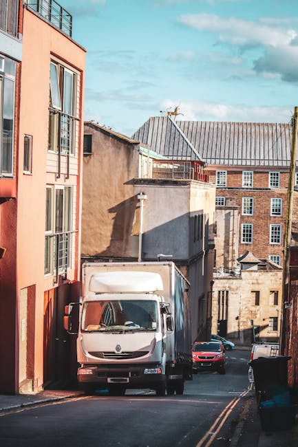 A narrow cobbled street with a curved layout, showing two white moving trucks parked side by side on the street surface. The trucks are positioned parallel to a brick retaining wall on the right, whic